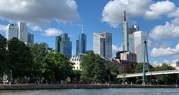 Skyline Frankfurt mit blauem Himmel, weißen Wolken im Hintergrund, Mainufer und Main im Vordergrund, Holbeinsteg am rechten Bildrand angeschnitten zu sehen.